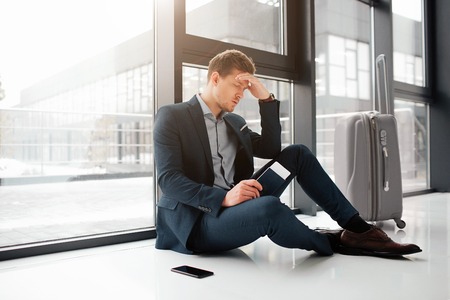 Young man has headache. He sit on floor at window in airport hall. Guy hold ticket with passport in hand. Phone and suitcase are besides.の写真素材