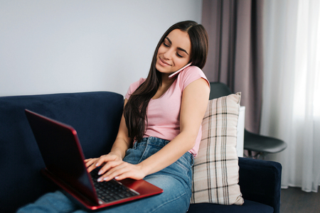Cheerful young woman work at home. She sit on sofa and talk on phone. Also model type on keyboard. She smiles.の写真素材