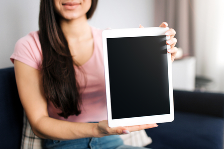 Cut view of young woman sit on sofa. She hold white tablet in hands and show it black screen to camera. Model smiles.の写真素材