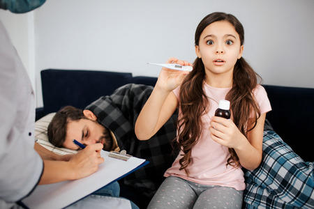 Scared small girl look on camera. She hold thermometer and bottle of syrup in hands. Female doctor...