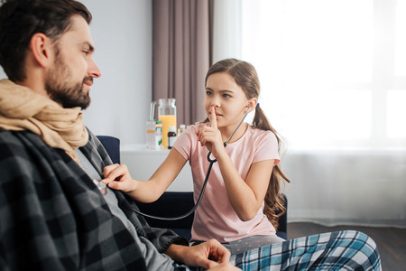 Small girl listen to dad's breathing through stethoscope. She hold finger on lips and look at him. Sick young man covered with blanket and scarfの写真素材