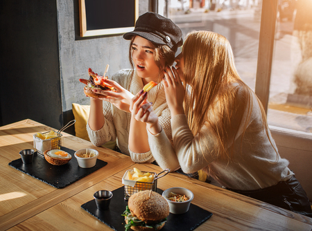 Beautiful young women sit in side at table. They gossip. Blodne girl wisper on ear to another girl. Model in cap is amazed. She hold burger.の写真素材