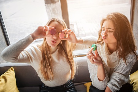 Playful young women eat macarons and cover eyes with it. They have fun together
