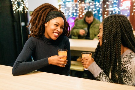 Two young african women sit in cafe. They have conversation. Model smile. They hold glasses of orange juiceの写真素材