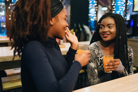 Cheerful african women sit in cafe and talk. They smile to each other. Models keep glasses with juice in handsの写真素材