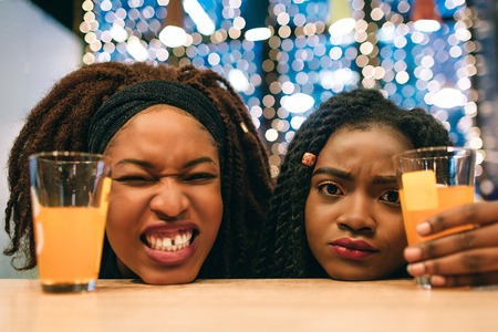 Funny young african women keep head on table. First model shrinks. Second one looks sad. She hold glass of juice with handの写真素材