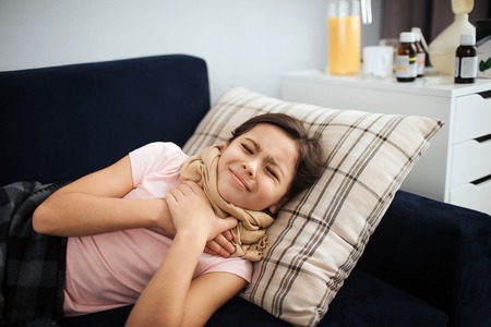 Sick girl lying on couch in room alone. She hold hands on throat. Girl suffer from pain. She shrinks...