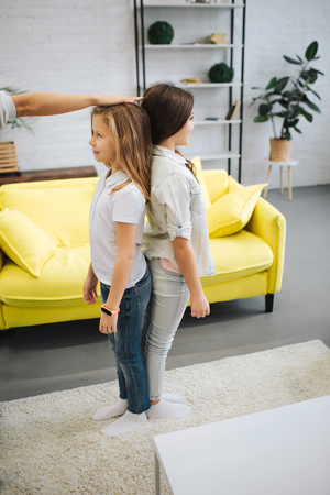 Two happy teenagers stand back to back in room and smile. Adult hol hand on girls head. He measure her growth. Brunette girl looks taller.の写真素材