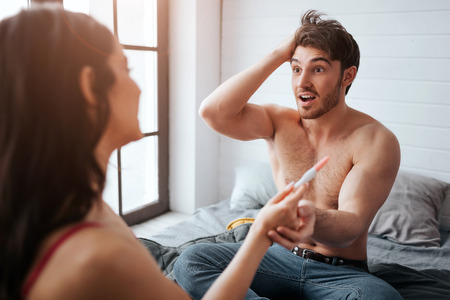 Amazed young man look at woman and hold her hand with pregnancy test. He is happy. They sit on bed. Happy woman look at him.の写真素材