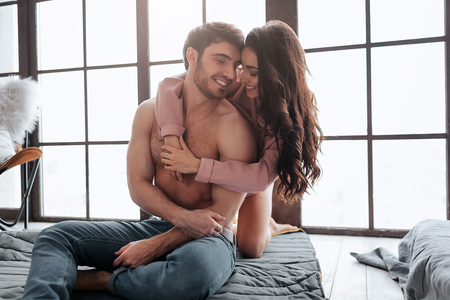 Happy and positive young couple sit together on floor in room. She stand behind and embrace man. He look at her and smile. Woman lean to guy.の写真素材