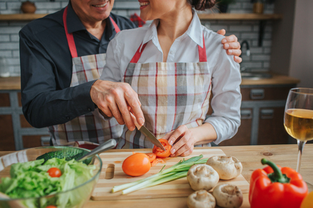 Cut view of man and woman stand together and cooking at table in kitchen. He has knife and cut tomato. She hold red vegetable with hands. They look at each other and smile.の写真素材