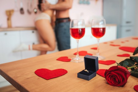 Cut view. Glasses of red wine stand on table with ring in box and rose. Young hot couple stand at kitchen cabinet and embraceの写真素材