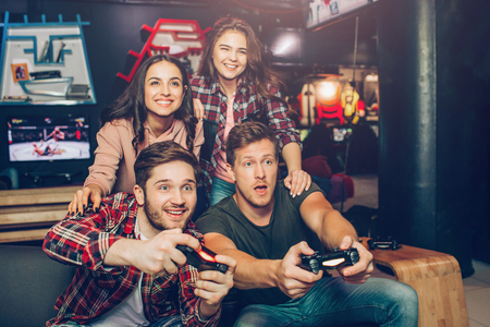 Excited young men sit on sofa in playing room. They hold joysticks and play game. Young women sit behind and cheer. People look stright forward.の写真素材
