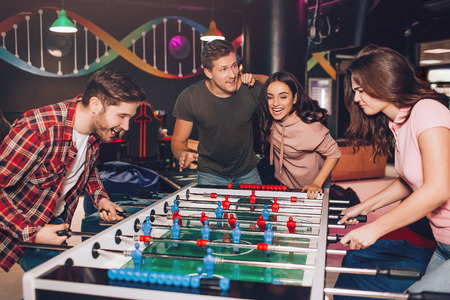 Enthusiastic excited young man and woman playing table soccer in room. Their friends stand beside...