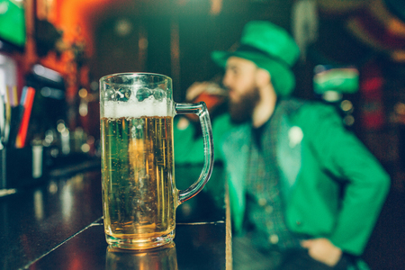 Serious concentrated young man in St. Patrick's suit sit at bar counter in pub alone. Mug of beer stand in front of him closer to cameraの写真素材