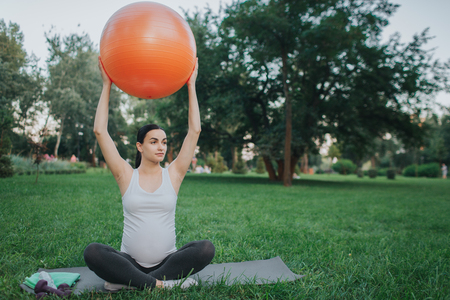Nice young pregnant woman sit in lotus pose in park. She hold big orange fitness ball with hands and look to right. She exercise.の写真素材