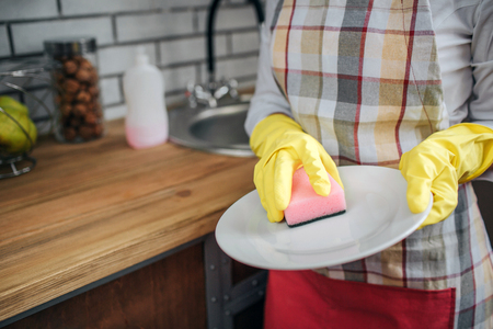 Close up of womans hands in yellow gloves washing dishes with sponge. She stand in kitchen at sink. Woman hold white plate.の写真素材