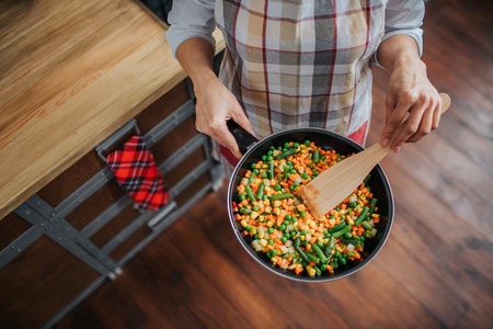 Close up of womans hands holding frying pan with food in it. She stand in kitchen. Woman wear apron.の写真素材