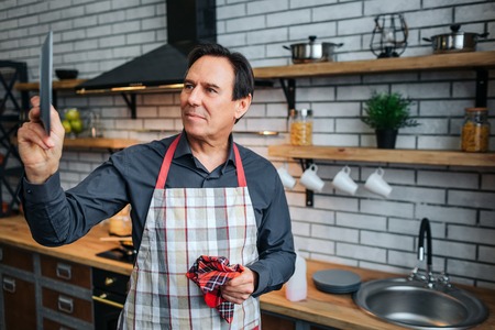 Serious man in apron stand in kitchen and look at plate in hand. He dry it. Also guy hold kitchen towel.の写真素材