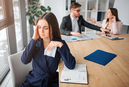 Sick young woman sit at table near window. She hold hand on head and suffer. Woman keep eyes closed. Her partners work behind.の写真素材