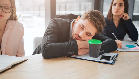 Tired young man sit at table and sleep in meeting room. He hold head on hands. Young women sit beside him and listen. They look concentrated.の写真素材