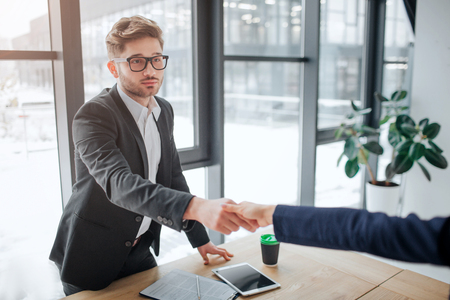 Serious young man stand at table in meeting room and shake woman hand. He look straight forward. Tablet with notes and cup of coffee on table.の写真素材