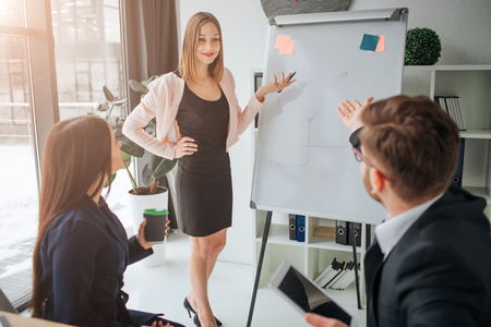 Confident young woman point on graphic on flipchart in room. She look at her colleagues and explain. Young man and woman sit at table and have questions.の写真素材