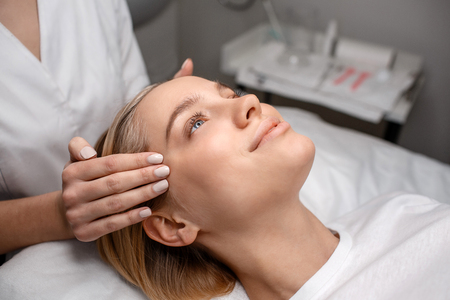 Nice young woman lying on couch in room. Beautician hands doing head massage. Model smile.の写真素材