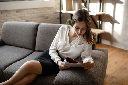Young thoughtful woman sit on sofa in office suit. She read and look down. Woman sit alone in room.の写真素材