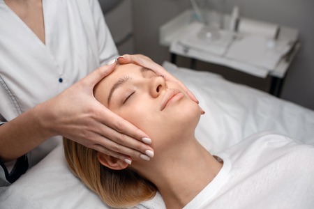 Pretty young woman enjoy face massage on couch. Beautisian touch it with bare hands. She stand at...