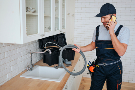 Young plumber talking on phone in kitchen at sink. He hold hose. Reparing. Toolbox on desk. Daylight.の写真素材