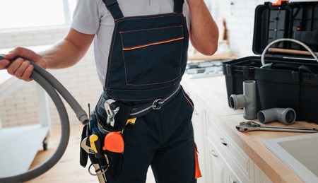 Cut view of man standing in kitchen and holding hose. He wear working uniform. Tools on belt. Guy stand at sink.の写真素材