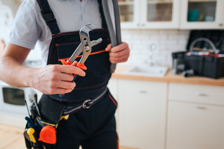 Cut view of man in working uniform hold wrench. He stand in kitchen. Daylight. Tools on belt. Toolbox on desk.の写真素材