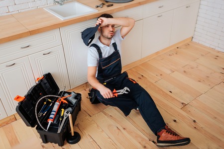 Tired exhausted plumber sit on floor in kitchen. He hold hand on forehead. Toolbox with wrech and other instruments stand on tableの写真素材