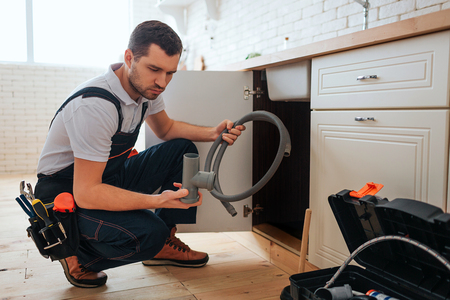 Serious concentrated young worker sit in squad pose in kitchen. He hold piece of pipe and hope. Guy doing repair. Opened toolbox on floor.の写真素材