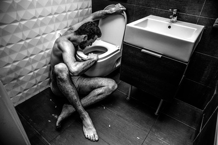 Colorless grey picture of sick young pale man sit on floor in rest room at toilet. He look inside it. Guy is going to burp. Bed feelings and terrible problems.の写真素材