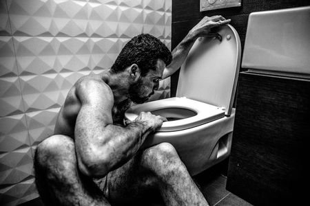 Young man sit at toilet and burp into it. Sickness and weak. He sit in cold rest room alone. Exhaustion and bad reaction. Black and white picture. Colorless.の写真素材