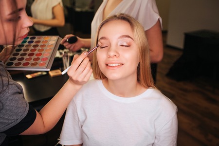 Nice young woman sit in beauty room with closed eyes. She smile. Make up artist put eyeshadows on eyeline. She hold palette in hands. Another profesional stand behind.の写真素材