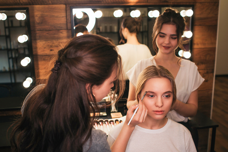 Serious blonde woman sit on chair in beauty room. Make up artist put some color on brows. Hairdresser sstand behind and lift hair up. She smile.の写真素材