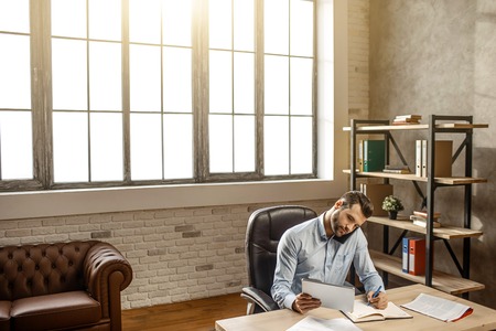 Young handsome businessman sit at table and writing in his own office. He talk on phone and look on tablet in hand. Busy and concentrated. Business talk. Meeting on-line.の写真素材