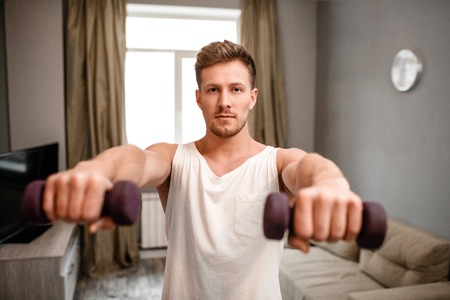 Young well-built man go in for sports in apartment. He hold dumbbells in front of himself and look on camera. Strong and muscular young man has workout.の写真素材