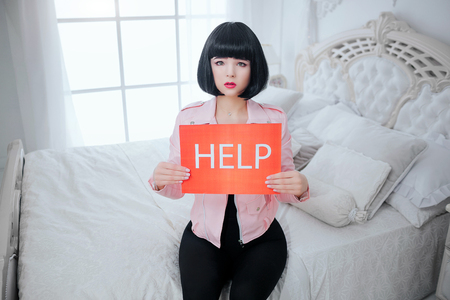 Glamour upset synthetic girl fake doll with short black hair is holding paper with word Help while sitting on the bed.の写真素材
