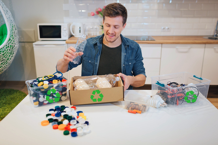 Recycling. Young smiling man putting emty plastic bottle into paper box while sitting at the table...