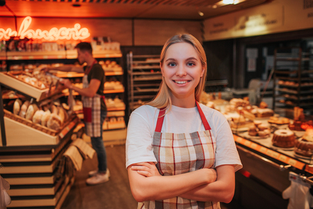 Happy young woman stand in grocery store. She pose nad look on camera. Happy worker smiling. Young man work behind. He put bread on shelf.の写真素材
