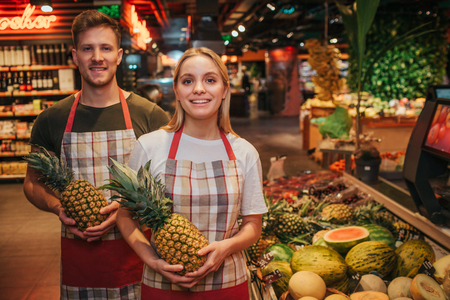 Young man and woman stand at fruit boxes in grocery store. They hold pineapples in hands and pose. Positive happy people workers.の写真素材