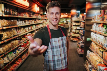 Handsome young man stand in grocery store among pasta shelfs. He reach hand to camera and smileの写真素材