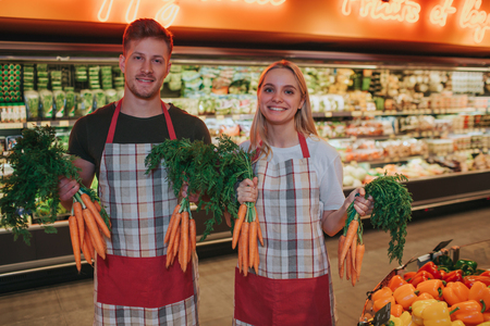 Young man and woman stand at vegetable basket in grocery store. They hold good healthy carrot in hands. Workers pose and smile.の写真素材