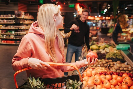 Young couple in grocery store. Woman hold pomegranat in hand and look at guy. He has grapes in hands. Smiling to each other.の写真素材