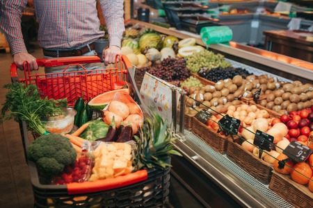 Young man alone in grocery store. Cut view guy hold trolley hand. Basket full of vegetables and fruit. Standing at baskets with good grocery.