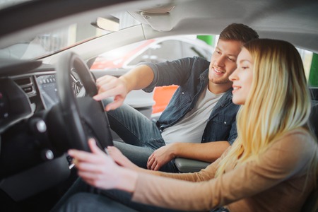 Young family buying first electric car in the showroom. Attractive smiling couple looking at buttons...